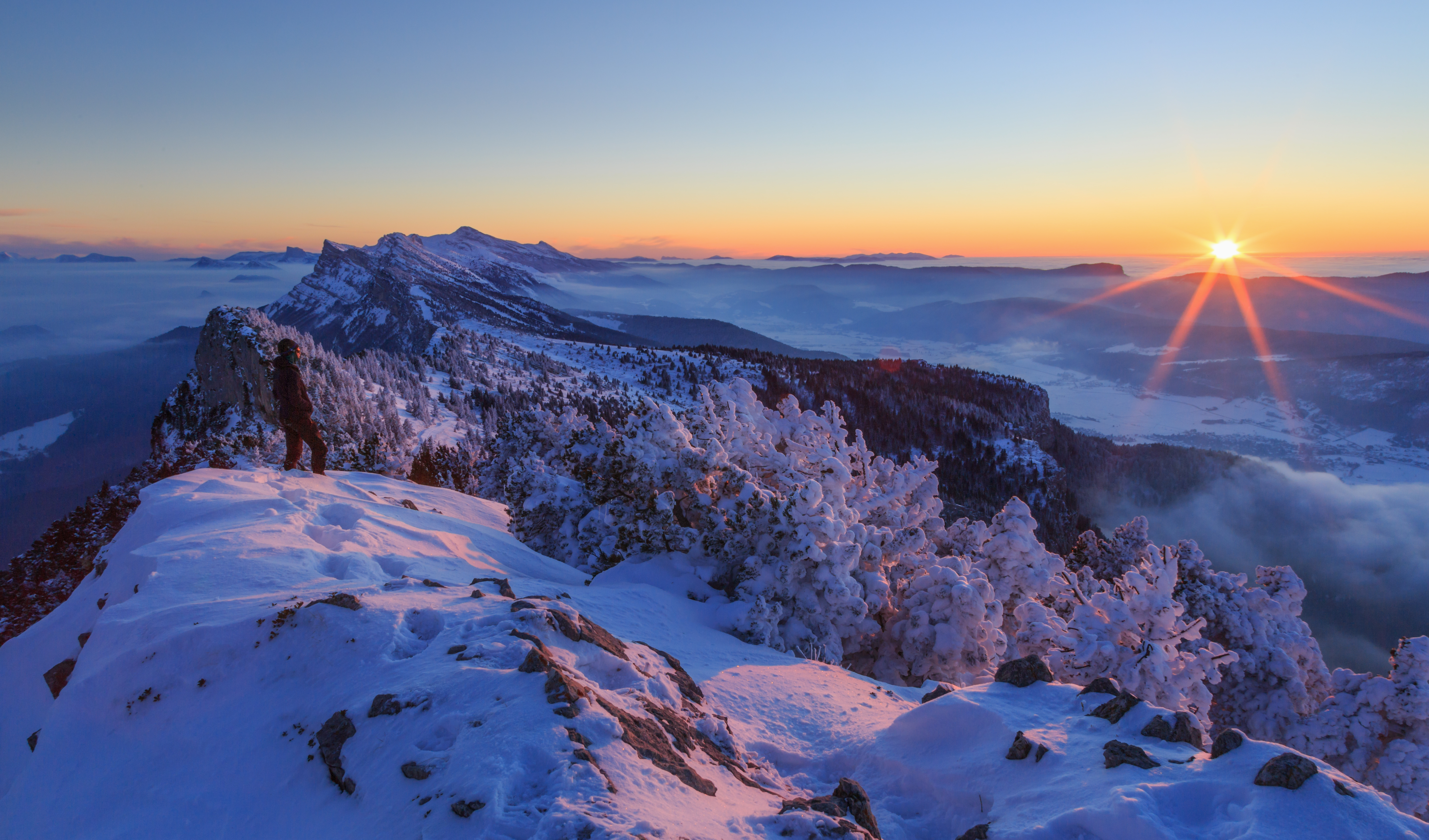 Hiker looking over a snow covered mountain range, Vercors, France, during a winter sunset.